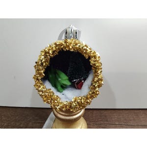 Pode incluir: Um enfeite de Natal esférico com uma borda dourada texturizada em torno de um interior oco. Dentro, uma figura de dinossauro verde está sobre uma base de neve branca ao lado de um pequeno pinheiro verde-escuro e uma pequena caixa de presente vermelha.