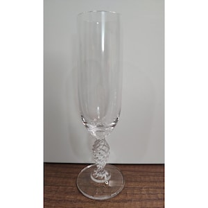 May include: A clear champagne flute with a long, slender bowl and a decorative, twisted stem. The base is round and clear. The glass is set against a neutral background and a wooden surface.