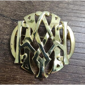 Könnte beinhalten: Goldfarbene Brosche mit einem komplizierten Design aus verschlungenen Buchstaben in einem kreisförmigen Rahmen. Die Brosche hat eine Nadel auf der Rückseite zur Befestigung. Das Design ist aufwendig und detailliert, mit poliertem Finish.