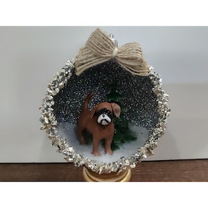 Könnte beinhalten: Eine runde, hohle Weihnachtskugel mit einer braun-schwarzen Hundefigur vor einem kleinen grünen Tannenbaum. Das Innere ist mit silbernem Glitzer ausgekleidet und der äußere Rand ist mit Silberfolie verziert. Oben befindet sich eine Schleife aus Jute.