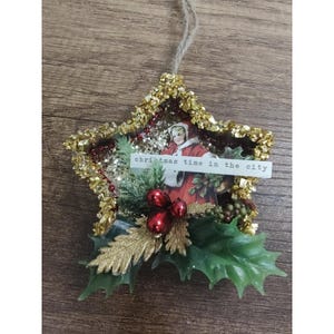May include: A star-shaped Christmas ornament featuring a gold glitter border, a vintage illustration of a person in a red coat, green holly leaves, red berries, and a white paper strip with the printed text "christmas time in the city".