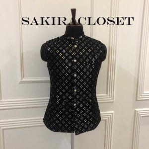 Puede incluir: Chaleco negro sin mangas con un patrón de diamantes de lentejuelas plateadas. El chaleco tiene cuello mandarín y se abrocha en la parte delantera con botones plateados. El texto "SAKIRA CLOSET" se muestra en la parte superior de la imagen.