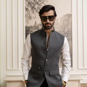 Peut inclure: Un homme porte un gilet gris anthracite avec un col officier sur une chemise blanche. Le gilet a deux poches avant et une fermeture boutonnée. Il porte des lunettes de soleil noires avec une étoile et une montre en or.
