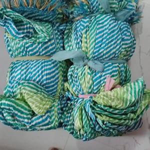 K&ouml;nnte beinhalten: Mehrere Stoffb&uuml;ndel in Blau- und Gr&uuml;nt&ouml;nen mit wei&szlig;en Streifen und Fransen. Das Gewebe ist mit B&auml;ndern gebunden. Die B&uuml;ndel liegen auf einer wei&szlig;en Oberfl&auml;che.