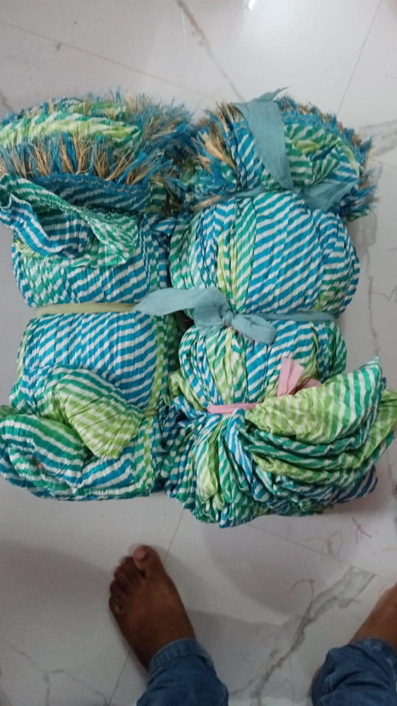 K&ouml;nnte beinhalten: Mehrere Stoffb&uuml;ndel in Blau- und Gr&uuml;nt&ouml;nen mit wei&szlig;en Streifen und Fransen. Das Gewebe ist mit B&auml;ndern gebunden. Die B&uuml;ndel liegen auf einer wei&szlig;en Oberfl&auml;che.