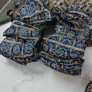 K&ouml;nnte beinhalten: Mehrere Stapel blauen Stoffes mit einem sich wiederholenden Muster aus wei&szlig;en und gelben Punkten und einem braunen Rand. Der Stoff ist mit gr&uuml;nen Gummib&auml;ndern gebunden und hat einen Fransenrand. Weitere Stoffe sind im Hintergrund zu sehen.