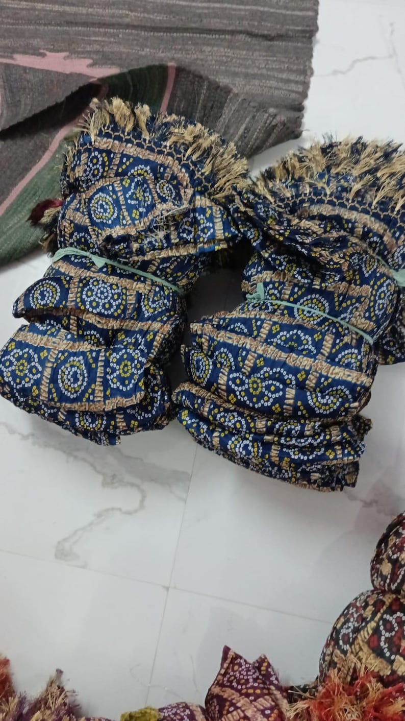 K&ouml;nnte beinhalten: Mehrere Stapel blauen Stoffes mit einem sich wiederholenden Muster aus wei&szlig;en und gelben Punkten und einem braunen Rand. Der Stoff ist mit gr&uuml;nen Gummib&auml;ndern gebunden und hat einen Fransenrand. Weitere Stoffe sind im Hintergrund zu sehen.