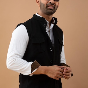Peut inclure: Un gilet noir avec un col officier et deux poches avant. Le gilet est porté sur une chemise blanche à manches longues rayée avec des poignets marron. L'ensemble est complété par des lunettes de soleil noires.