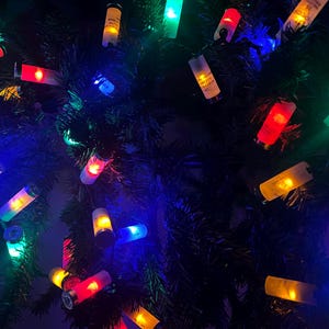 Puede incluir: Una cadena de luces navideñas con bombillas multicolores, incluyendo rojo, verde, azul y amarillo. Las luces están enhebradas en un árbol de Navidad artificial verde oscuro.