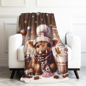 May include: A cozy throw blanket featuring a Highland cow calf wearing a winter hat and scarf. The blanket has a brown background with snowflakes and Christmas ornaments. A glass of hot chocolate with whipped cream and cinnamon sticks is also visible.