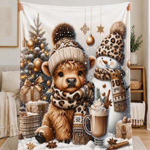 May include: A white fleece blanket featuring a fluffy brown Highland cow wearing a leopard print scarf and beanie, sitting next to a snowman in a matching leopard print hat and scarf, a glass of hot cocoa, Christmas tree, and gift boxes.