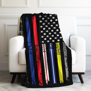 May include: A black fleece throw blanket with a thin red, white, and blue stripe down the center. The stripe is flanked by thin stripes of blue, red, yellow, and gray. The text "Corrections", "Dispatch", "EMS", "Nurse", "Firefighter", and "Police" is printed vertically on the stripes. The blanket is folded and draped over a white armchair.