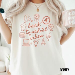 Puede incluir: Una camiseta de color marfil con un estampado gráfico rojo con el texto back to school vibes rodeado de ilustraciones de una mochila, un birrete, una calculadora, un portátil, una bombilla, un compás, un globo terráqueo y un tubo de ensayo.