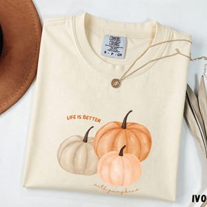 Puede incluir: Una camiseta de color marfil con un gráfico de tres calabazas en tonos naranja, melocotón y beige. El texto sobre las calabazas dice "LIFE IS BETTER" y el texto debajo dice "with pumpkins". La camiseta tiene cuello redondo y una etiqueta.