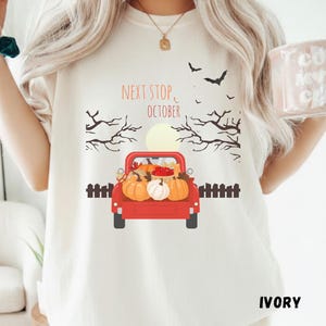Puede incluir: Una camiseta de color marfil con un gráfico de una camioneta roja llena de calabazas. Sobre la camioneta, el texto dice NEXT STOP, OCTOBER. El diseño incluye siluetas de árboles desnudos, una luna llena y murciélagos volando.