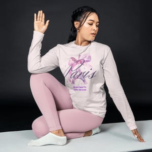 Puede incluir: Una mujer lleva una camiseta rosa de manga larga con un diseño de acuarela de la Torre Eiffel y la palabra "París" en escritura morada. La camiseta también tiene el texto "Where hearts unite forever". Lleva pantalones de yoga rosas y calcetines blancos.