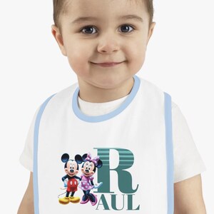 Puede incluir: Babero blanco para bebé con ribete azul que presenta a Mickey y Minnie Mouse, una letra R verde y el nombre "Paul".