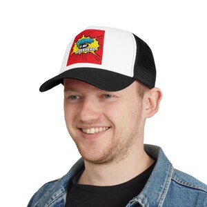 Puede incluir: Una gorra de camionero negra y blanca con un gráfico de estilo cómic rojo, amarillo y azul que dice "Trump is my Superhero".