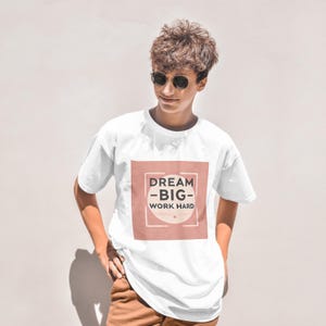 Puede incluir: Camiseta blanca con un gráfico cuadrado rosa que dice "Dream -Big- Work Hard".