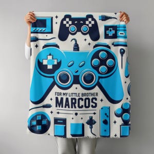 Puede incluir: Una manta de color claro con un diseño de mando de videojuegos azul y blanco. La manta tiene un gran mando central con el texto "FOR MY LITTLE BROTHER MARCOS". Gráficos adicionales con temática de juegos están dispersos por toda la manta.