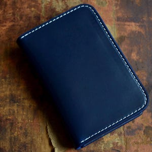Puede incluir: Una cartera de cuero azul marino oscuro con esquinas redondeadas y costuras blancas en contraste a lo largo de los bordes. La cartera tiene una textura de superficie lisa y mate y una forma rectangular con un pliegue en el lado izquierdo.