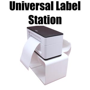 Puede incluir: Una impresora de etiquetas blanca y negra con el texto "Universal Label Station" impreso arriba. La impresora dispensa un rollo de papel de etiquetas blanco. La impresora se encuentra sobre un soporte blanco.