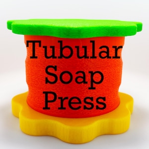 May include: A three-part soap press in bright colors. The base is yellow, the middle section is orange, and the top is green. The words "Tubular Soap Press" are printed in black on the orange section.
