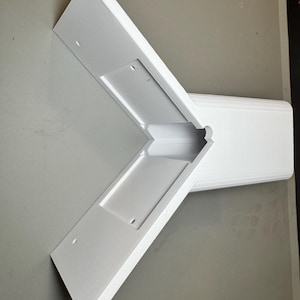 May include: A white, angular, three-dimensional object with a cross-like shape. The object has a rectangular base and two arms extending at a 90-degree angle. Each arm has a rectangular indentation with two small holes. The object appears to be made of a solid, matte material.