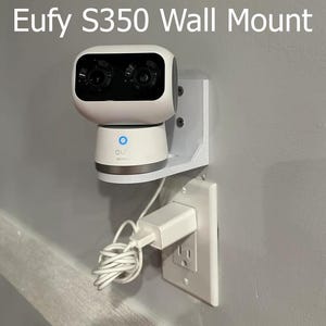 May include: A white Eufy S350 security camera mounted on a white wall mount. The camera is plugged into a white electrical outlet with a white cord.
