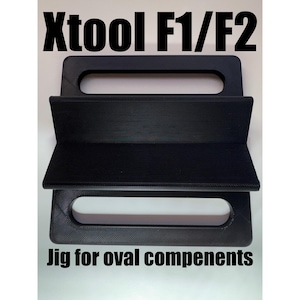 May include: Black jig for oval components with the text "Xtool F1/F2". The jig has a handle and a shelf. The text "Jig for oval components" is printed on the bottom of the jig.