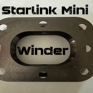 May include: A black, rectangular Starlink Mini winder with rounded corners and a large central oval cutout. The object features eight circular holes arranged around the perimeter. The text Starlink Mini and winder are printed on the surface.