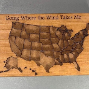 May include: Wooden plaque featuring a detailed engraving of the United States map. The phrase "Going Where the Wind Takes Me" is etched above the map. The wood has a warm, natural tone, and the engraving is a darker brown.