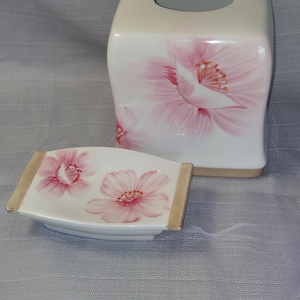Puede incluir: Un cubridor de caja de pañuelos de cerámica blanca y un jabonera a juego con diseños florales rosas. El cubridor de caja de pañuelos tiene forma cuadrada con una parte superior redondeada y una ranura para pañuelos. La jabonera es rectangular con bordes redondeados. Ambas piezas tienen un borde dorado.