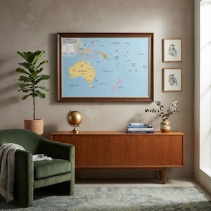 May include: A large framed map of Australia and Oceania with a light blue background and yellow landmasses hangs above a wooden sideboard. The room includes a green armchair, a potted fiddle leaf fig, a globe, and two small bird prints.