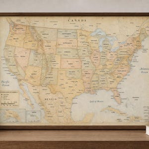 May include: A framed wall map of the United States of America featuring a vintage-style aesthetic with muted tan, beige, and soft green color tones. The map displays state boundaries, major cities, and geographical features, with text labels for Canada, Mexico, and the Atlantic Ocean.