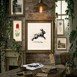 May include: A framed black and white ink illustration of a leaping hare centered on a wall. The artwork is displayed in a gold frame against a rustic brick wall, surrounded by hanging green plants and other framed botanical and landscape prints.