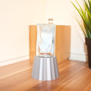 Könnte beinhalten: Eine klare Glasvase mit gedrehtem Design steht auf einem grauen, geometrischen Sockel. Die Vase wird auf einer Holzoberfläche ausgestellt, mit einer Topfpflanze im Hintergrund.