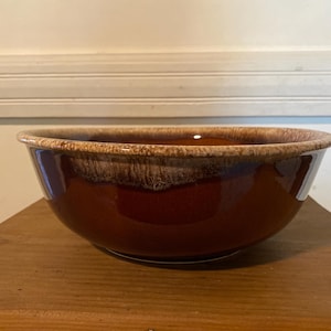 May include: A round ceramic bowl featuring a deep, glossy brown glaze on the lower portion and a lighter, textured beige and tan drip pattern along the upper rim. The bowl has a wide opening and a smooth, curved silhouette with a distinct colour transition.