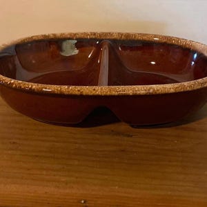 May include: An oval-shaped ceramic serving dish with a central divider. The interior features a deep, glossy brown glaze, while the outer rim is finished with a textured, speckled beige and tan pattern that contrasts with the smooth dark centre.