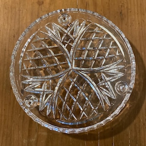 May include: A circular clear glass coaster featuring a faceted diamond-cut pattern radiating from the centre. The coaster has a serrated edge and three small, raised circular feet on the underside to provide stability and elevation on flat surfaces.