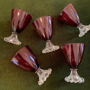 May include: Five ruby red stemmed glasses with clear, decorative bases. The glasses have a flared bowl shape and are arranged on a green surface. The glasses are likely for serving wine or other beverages.