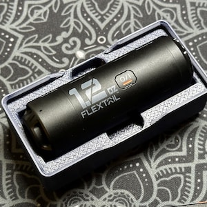 May include: A black flashlight with a silver label that reads "12 oz Flextail" and a small orange button. The flashlight is in a black plastic case.