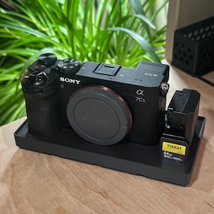 May include: A black Sony Alpha 7CR camera with a lens cap, resting on a black tray. A battery charger with a yellow and black label is next to the camera. The background includes green foliage.
