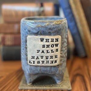 May include: A blue ceramic planter with a textured surface and a rectangular plaque that reads "WHEN SNOW FALLS NATURE LISTENS." The planter has a square base and is supported by four small feet. The background includes books.
