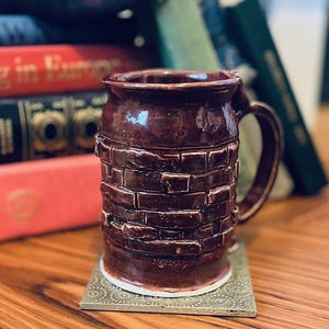 Puede incluir: Taza de cerámica burdeos oscuro hecha a mano con un diseño texturizado en forma de ladrillo. La taza tiene un asa grande y se apoya en un posavasos decorativo. Los libros son visibles en el fondo, lo que sugiere un tema literario y acogedor.