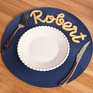 May include: A place setting with a navy blue woven placemat, a white plate, and silverware. The name "Robert" is spelled out in yellow yarn. A small potted plant sits on the wooden table.