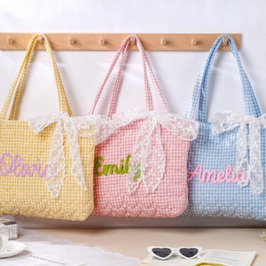 Op de afbeelding: Drie geruite draagtassen in geel, roze en blauw, elk met een naam geborduurd in een contrasterende kleur. De tassen hebben witte kanten accenten en bijpassende handvatten, hangend aan een houten rek.