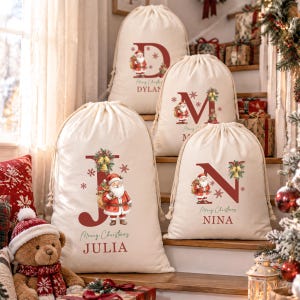 May include: Four off-white canvas drawstring gift bags featuring large red letters, illustrations of Santa Claus, gold bells, and snowflakes. The bags are personalized with the names Julia, Nina, Dylan, and an obscured name, with Merry Christmas text.