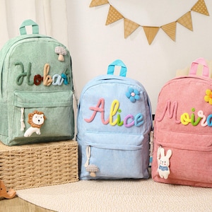 May include: Three corduroy backpacks in mint green, light blue, and pink. Each backpack has a name in colorful letters, a front pocket, and a top handle. The green backpack has a lion and mushroom, the blue has a flower, and the pink has a flower and a bunny.