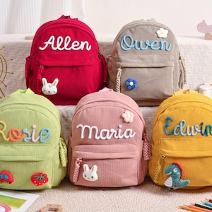 May include: Five corduroy backpacks in red, green, pink, gray, and yellow, each with a name embroidered on the front. The backpacks have a front pocket and a zipper closure. Each backpack has a decorative charm.